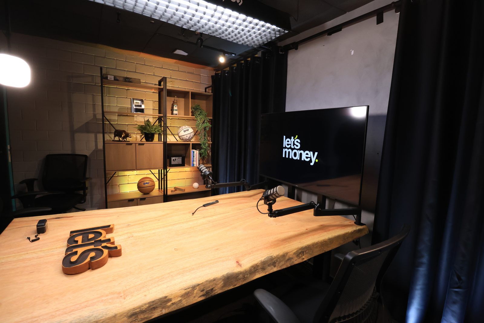 Set do estúdio com a mesa de madeira e o painel Let's Money ao fundo