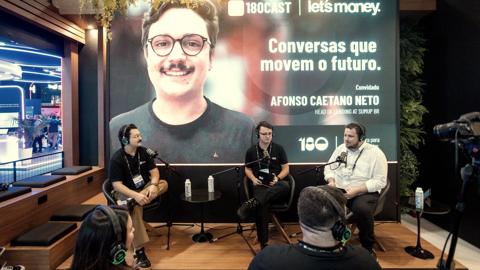 Palco do 180Cast na Febraban Tech 2026: três pessoas de fone ao microfone diante do telão com o logo da Let's Money