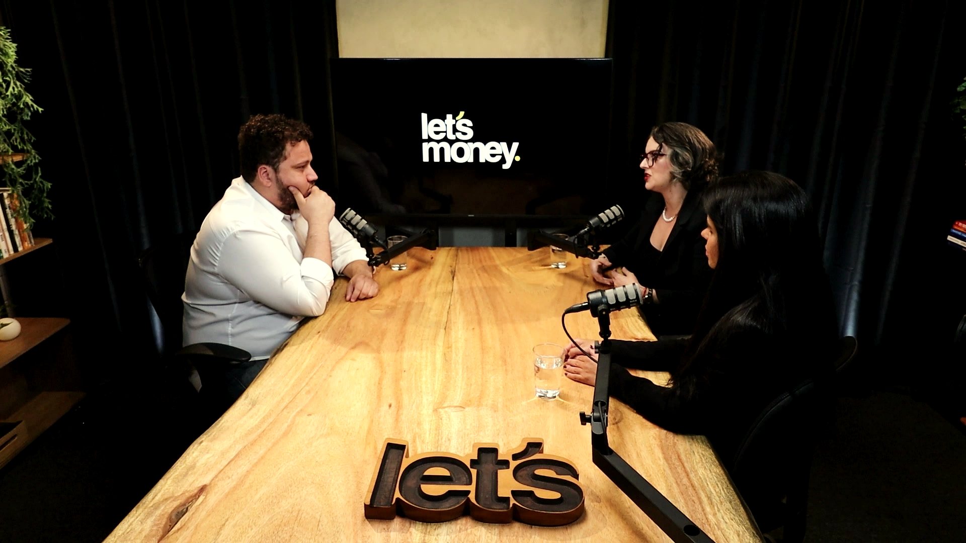 Gabriel Pereira e duas convidadas na mesa de madeira do estúdio, com o logo da Let's Money na tela ao fundo